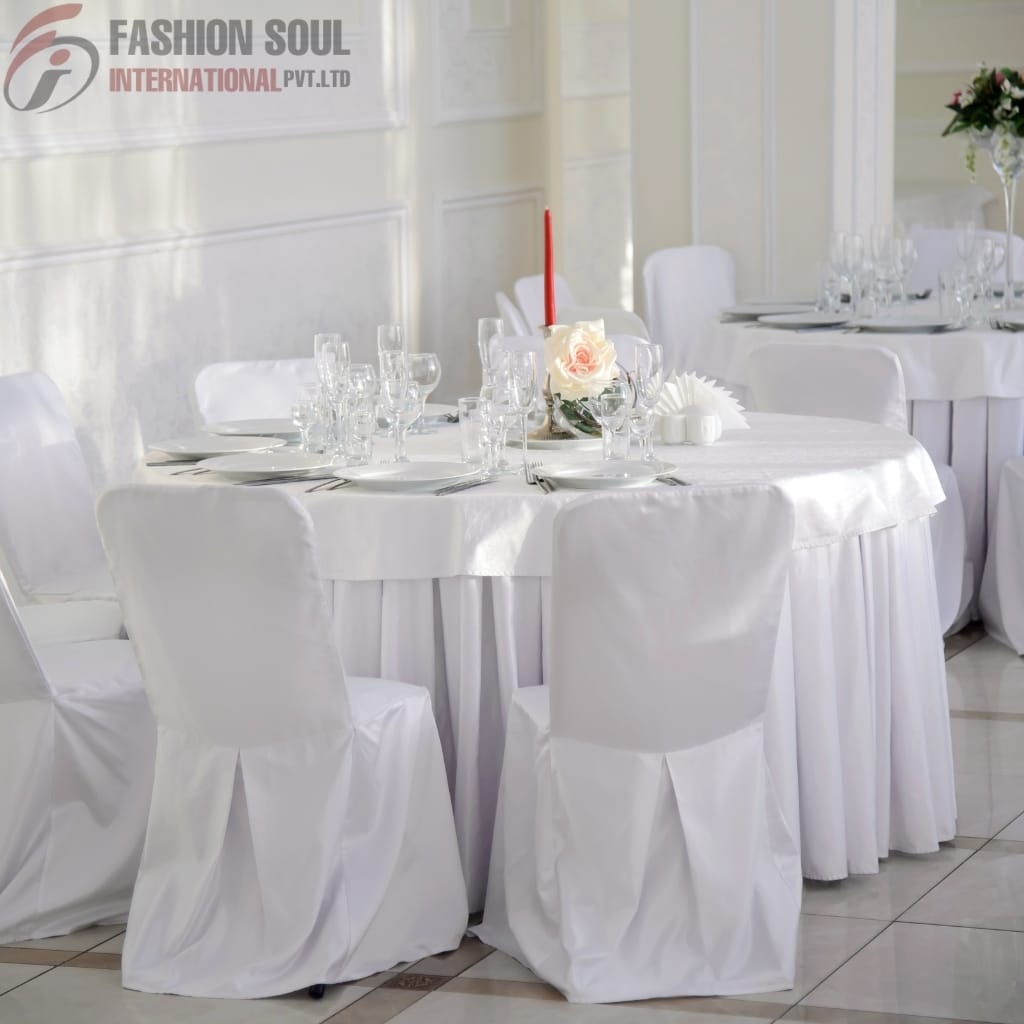 A round banquet table set for a formal event, covered with a white tablecloth and surrounded by white chairs with matching covers and tied with brown sashes. The table is elegantly decorated with wine glasses, plates, cutlery, a pink rose centerpiece, a tall red candle, and some decorative elements. The background shows a softly lit, white-walled room with elegant paneling.