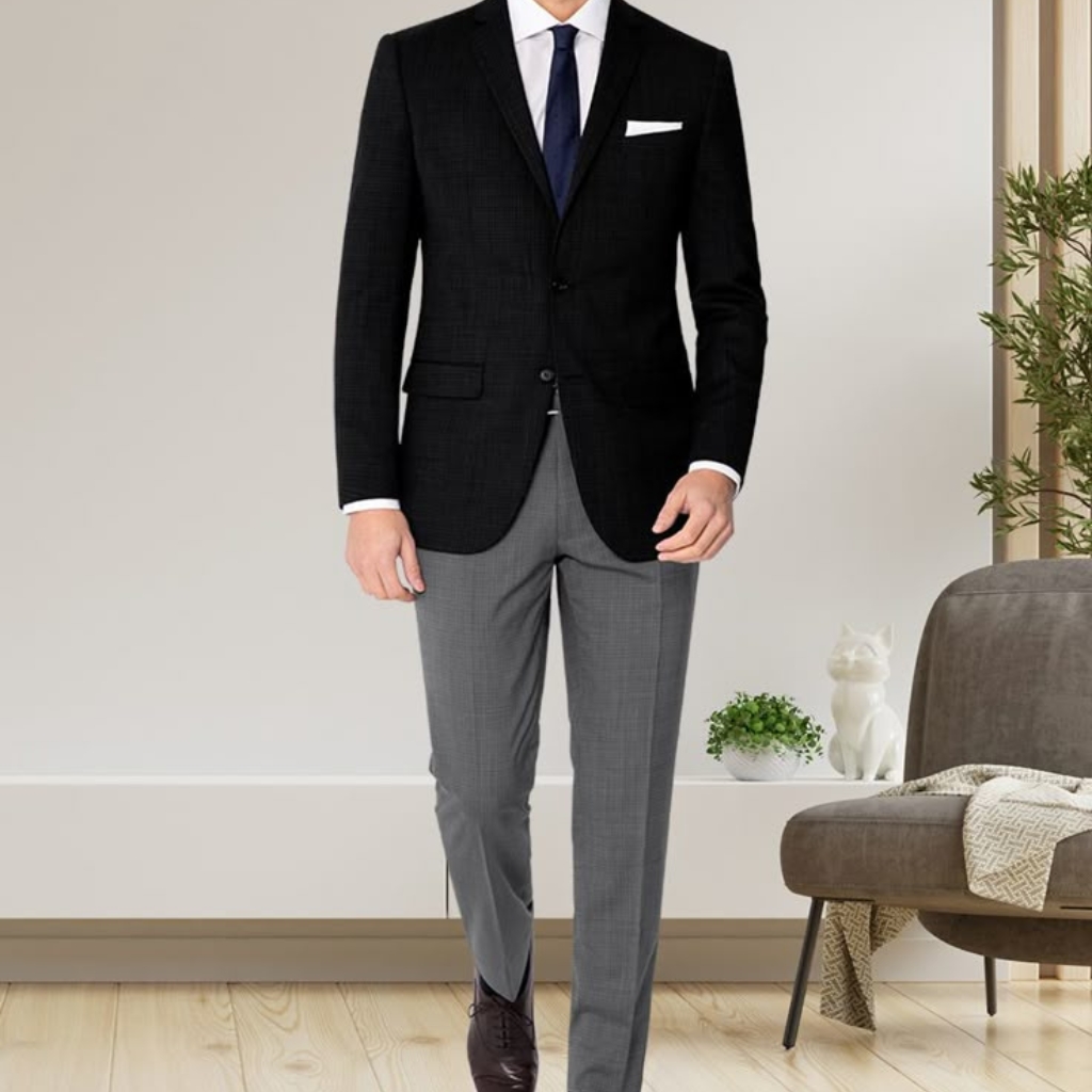 A man dressed in a formal black sport coat paired with light grey dress pants, white shirt, navy tie, and dark shoes, standing in a modern indoor setting.