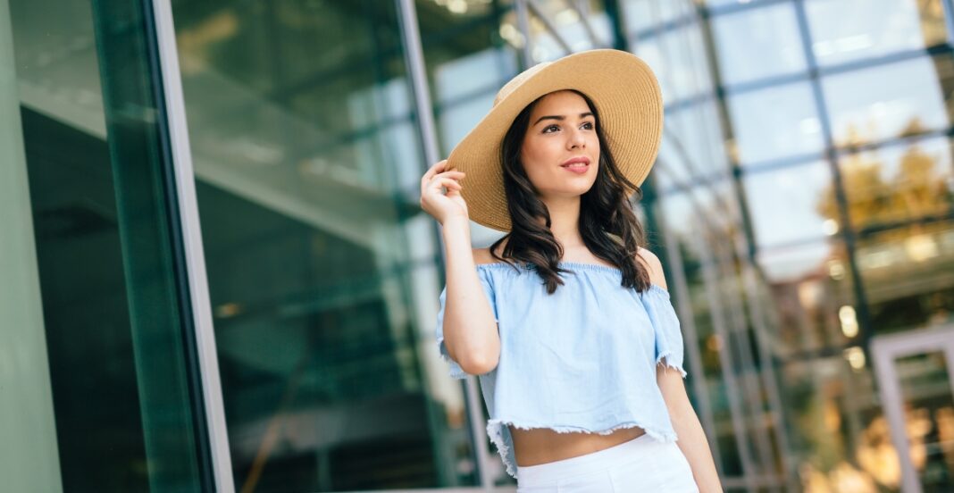 A young woman standing outdoors near a modern glass building, wearing a wide-brimmed hat and a light blue off-the-shoulder crop top.