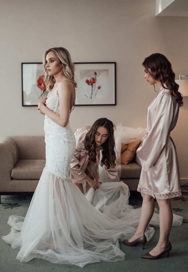 Three women are preparing for a wedding in a cozy room. One stands in a white mermaid wedding gown with a sheer train, another in a satin robe, and the third is kneeling, adjusting the dress.