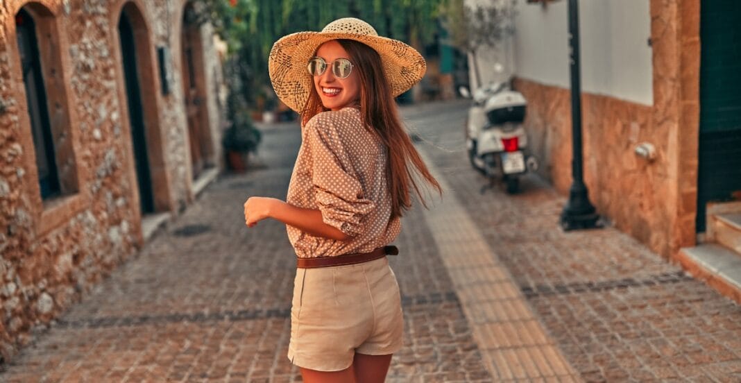 A young woman with long hair is smiling and looking over her shoulder while standing on a cobblestone street in a charming town. She is wearing a wide-brimmed straw hat, sunglasses, a polka-dot blouse, and beige shorts.