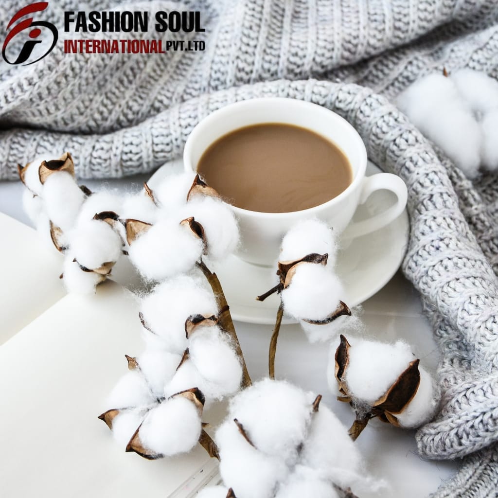 A white cup filled with coffee placed on a saucer, surrounded by cotton branches with fluffy cotton buffs. In the background, a knitted grey blanket and the logo of "Fashion Soul International Pvt Ltd" are visible.