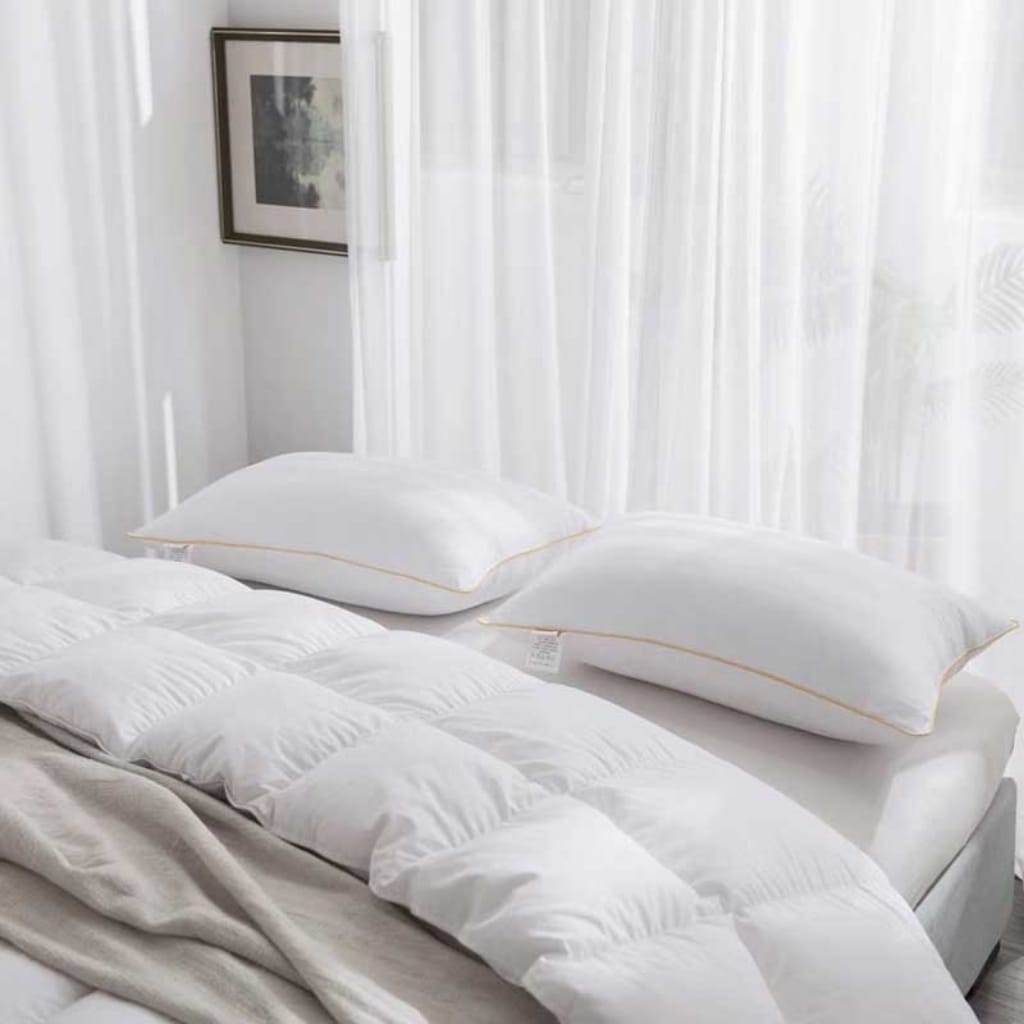 A neatly made bed with white pillows, a white comforter, and beige sheets, set against a background of sheer white curtains and a framed picture on the wall.