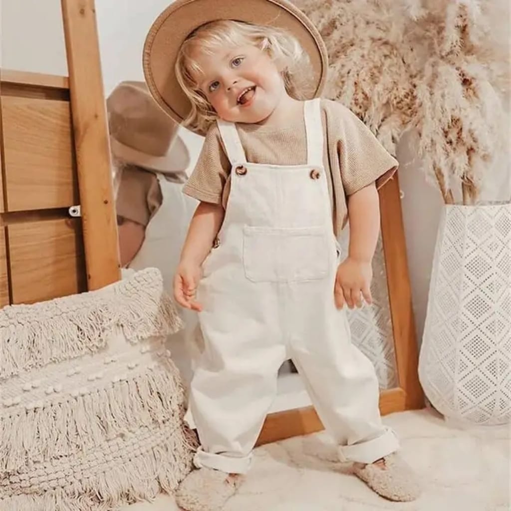 A joyful young child with blonde curly hair, wearing a beige hat, beige shirt, and white overalls, standing indoors next to a knitted pillow and tall vase with dried pampas grass.