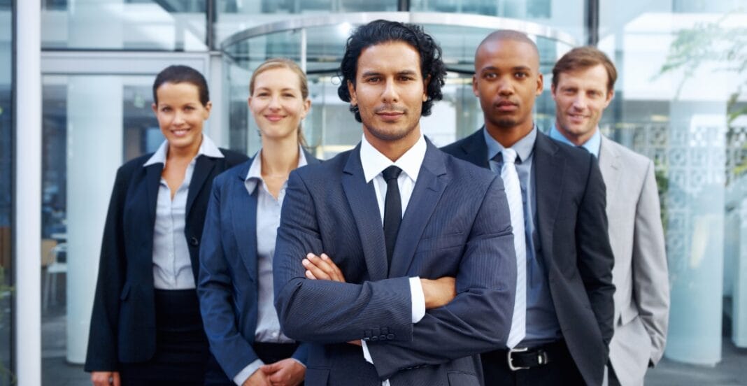 A group of five professionally dressed businesspeople standing outdoors in front of a modern office building, with a man in the foreground crossing his arms confidently.