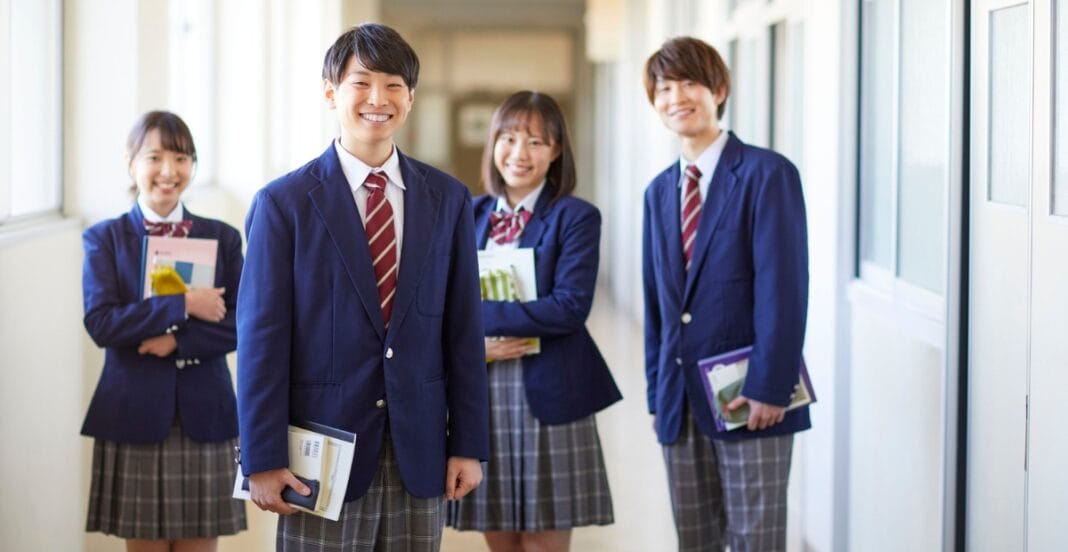 A group of four smiling students in school uniforms standing in a corridor, holding books and notebooks.