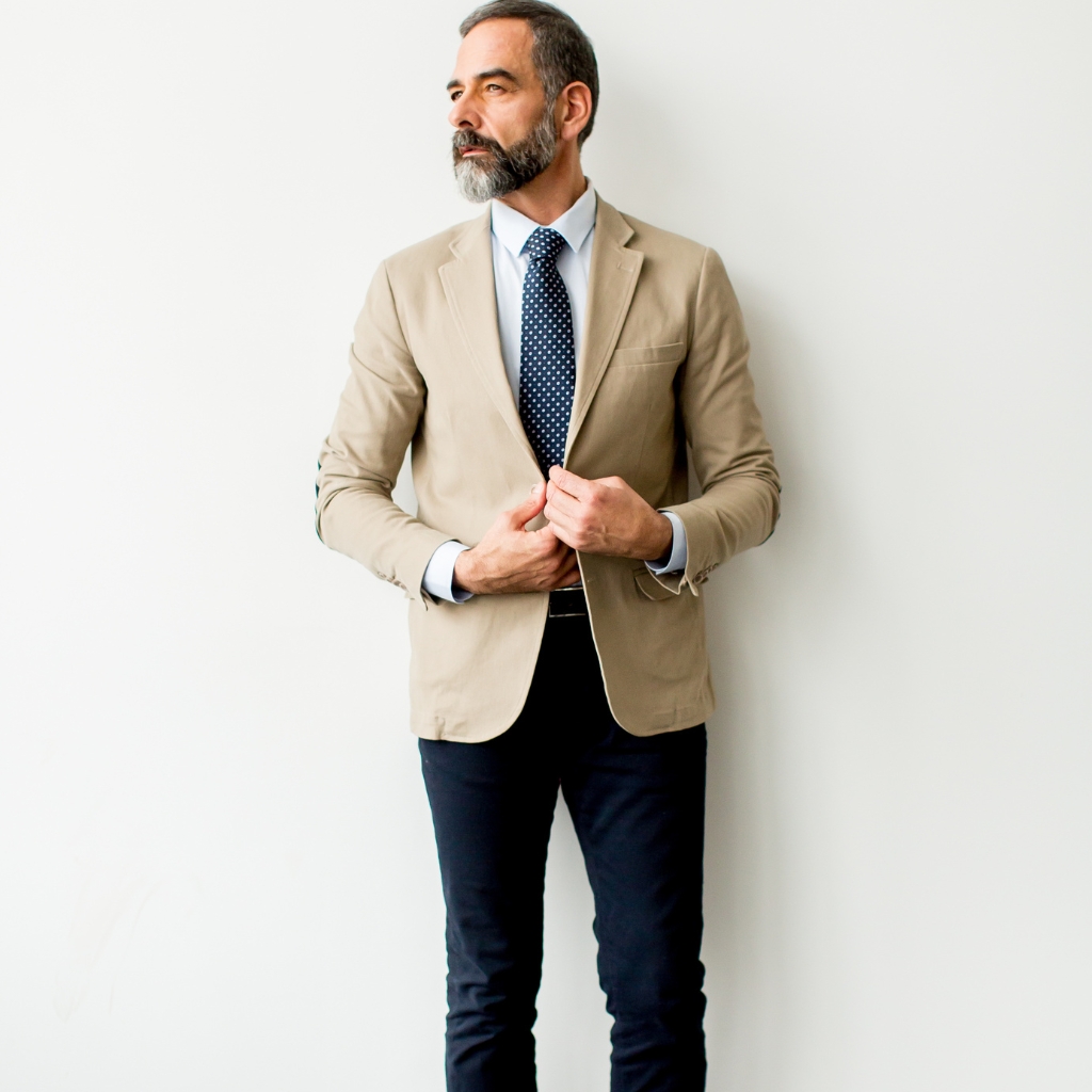 A man dressed in business casual attire, wearing a beige sport coat, a light blue shirt, a navy polka-dot tie, and dark jeans, standing against a plain white wall.