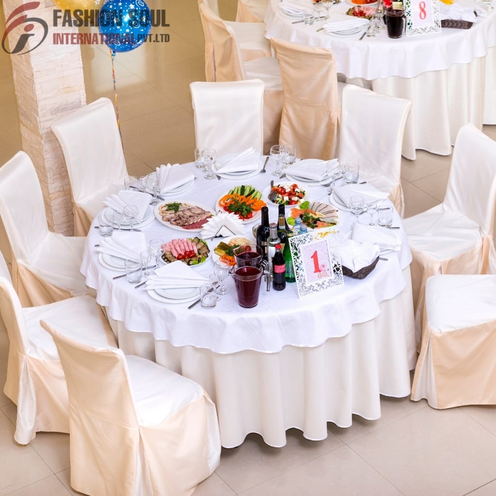 A round banquet table decorated with white tablecloth and elegant white chair covers, set with plates, glasses, cutlery, and various dishes. The table has a table number card with the number "1" and a centerpiece with beverages and condiments.