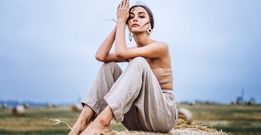 A young woman sitting on a haystack outdoors, with a calm expression, wearing beige pants, a strapless top, and large earrings, with a blurred field and blue sky in the background.