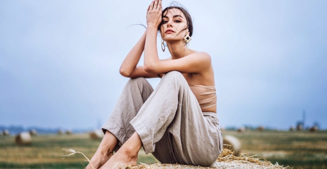 A young woman sitting on a haystack outdoors, with a calm expression, wearing beige pants, a strapless top, and large earrings, with a blurred field and blue sky in the background.