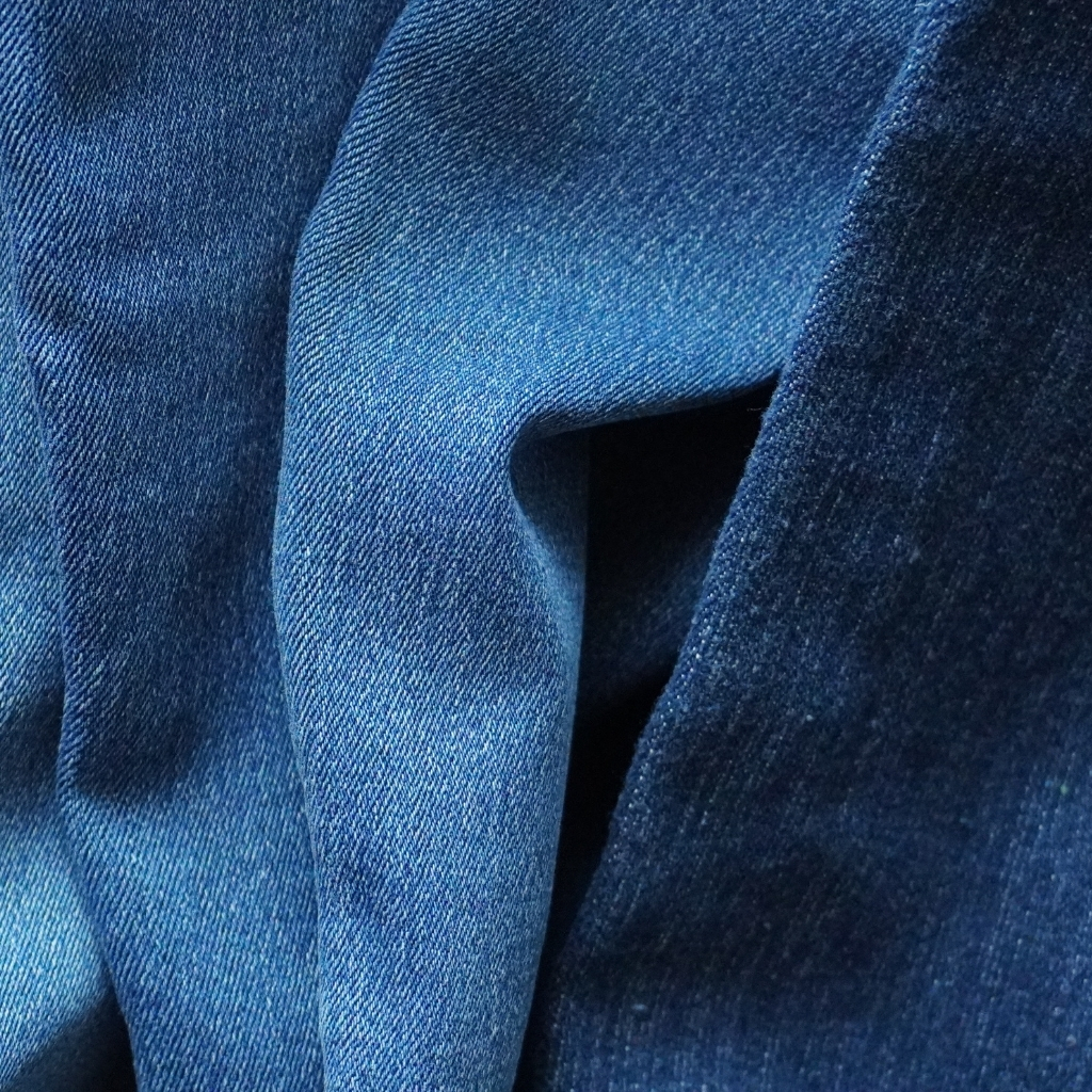 A close-up of dark blue denim fabric showing detailed texture and weave, with folds creating shadows and highlights.