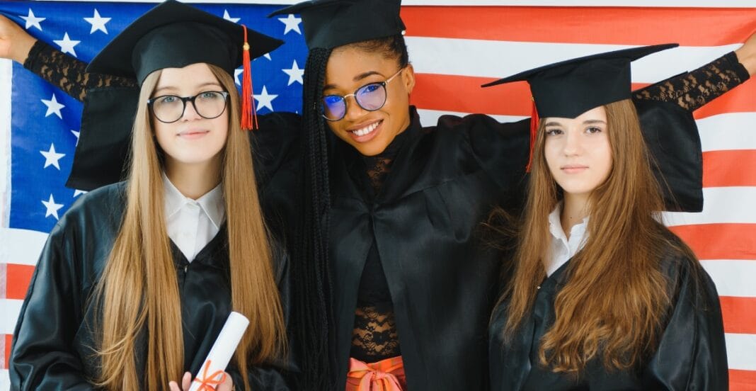 Three young women in graduation caps and gowns standing in front of an American flag, smiling and celebrating graduation.