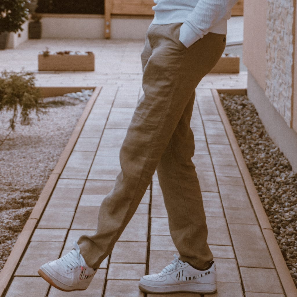A person standing on a wooden pathway outdoors, wearing beige pants, white sneakers with black stripes, and a light gray hoodie with hands in pockets. The face is not visible.