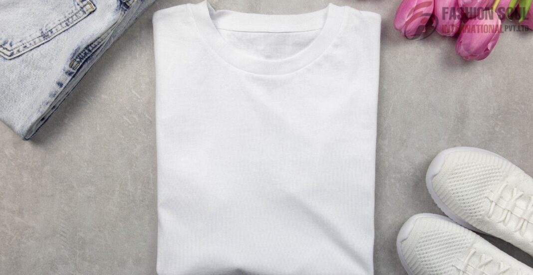A neatly folded plain white t-shirt placed on a light gray surface, with part of a pair of white sneakers, pink flowers, and a folded denim garment visible around it.