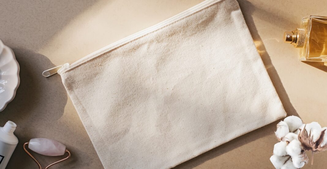 A beige fabric zippered pouch on a neutral surface, surrounded by a cotton flower, a perfume bottle, a cooling roller, and part of a container.