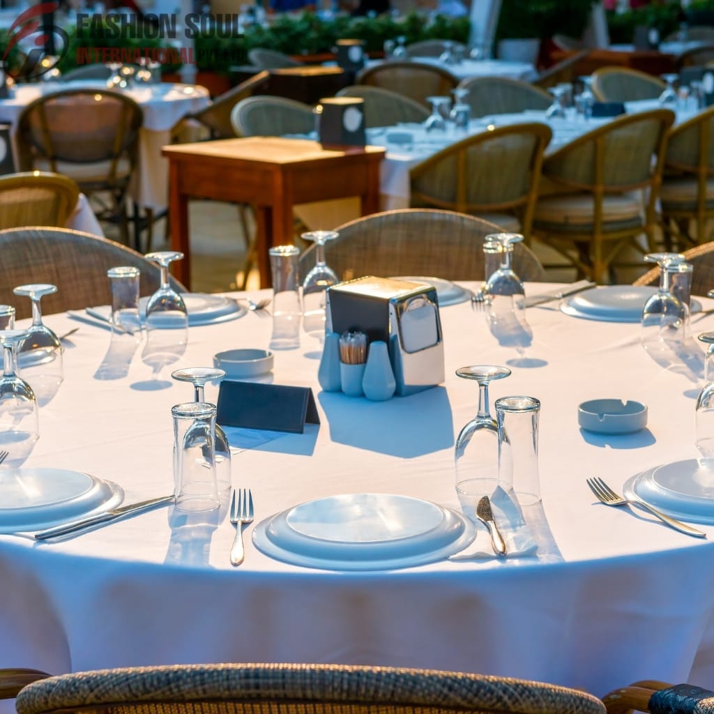 A round dining table set for a formal event with a white tablecloth, arranged with plates, glasses, silverware, and a centerpiece, surrounded by wicker chairs in an outdoor setting.