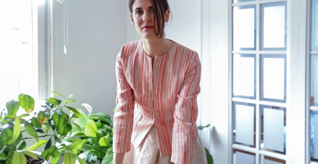 A woman with dark hair styled in a loose side part, wearing a light-colored, striped blouse, and leaning forward in an indoor setting with natural light, greenery, and modern furnishings.
