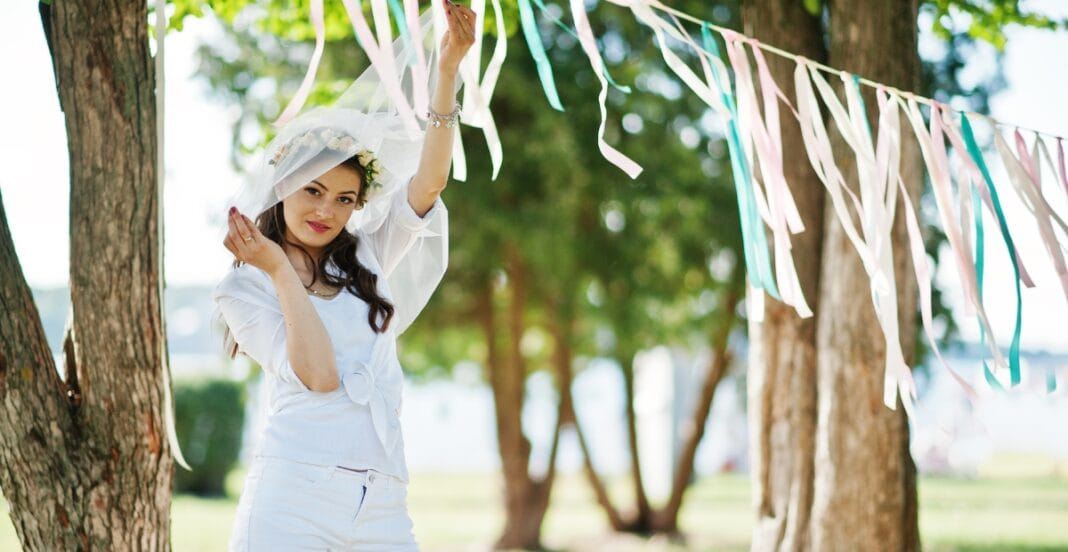 A young woman dressed in white with a veil and floral headband, standing outdoors among trees with colorful ribbons hanging above her, celebrating a special occasion.