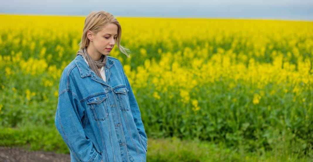 A young woman with blonde hair, wearing a denim jacket, stands in a field of yellow flowers with a contemplative expression.