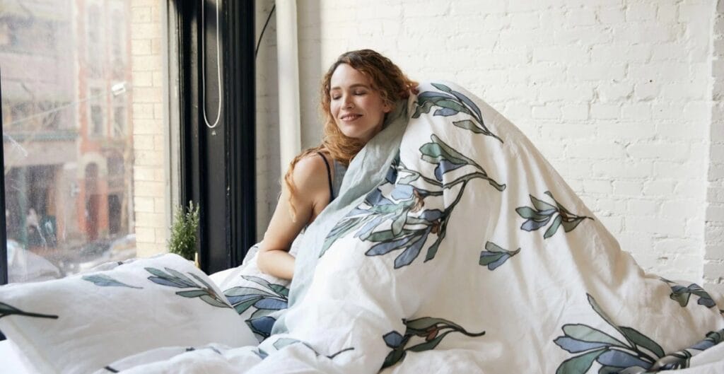 A woman with curly hair is sitting in bed near a large window, smiling and wrapped in a white blanket with a leafy pattern. She appears relaxed and content.