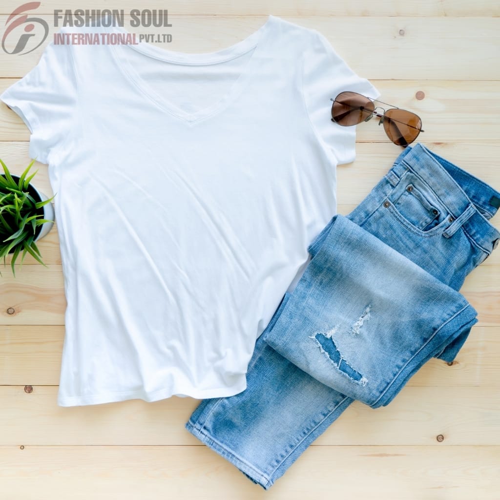 A flat lay of a casual outfit on a light wooden surface, including a plain white V-neck T-shirt, a pair of distressed blue jeans, a pair of brown sunglasses, and a small green potted plant.