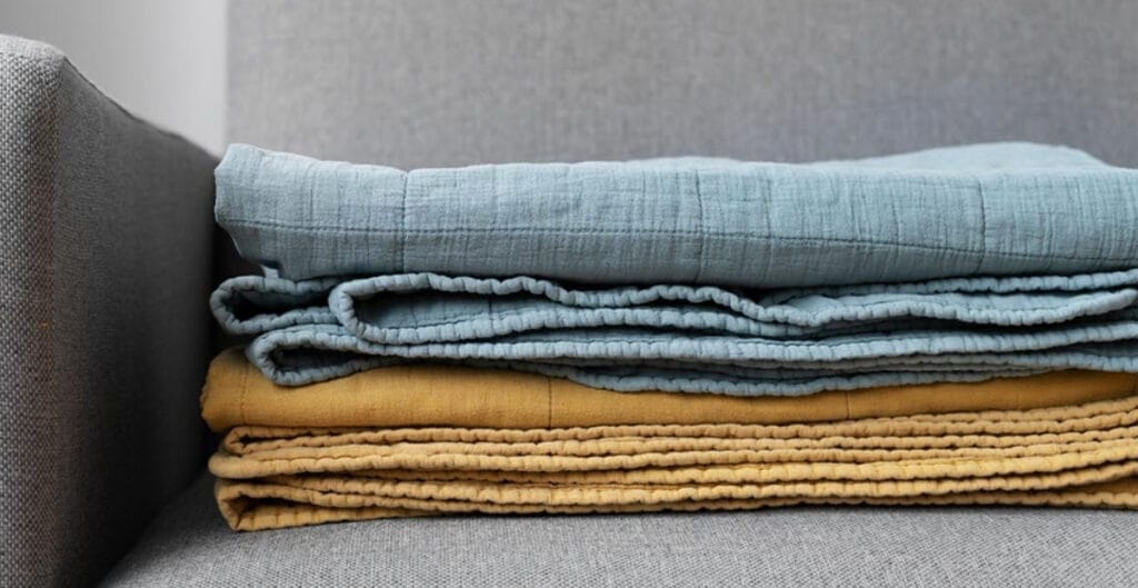 Three folded pieces of fabric in light blue, mustard yellow, and tan colors are stacked on a gray upholstered surface with a gray cushion behind them.