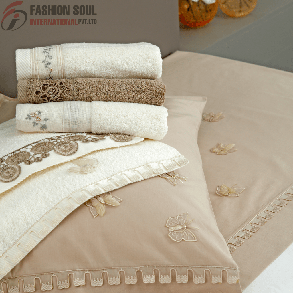 A neatly arranged display of folded embroidered and textured towels and bed linens in neutral tones with butterfly and floral embroidery, placed on a bed with pillows. The background features a logo of "Fashion Soul International Pvt. Ltd." in the upper left corner.