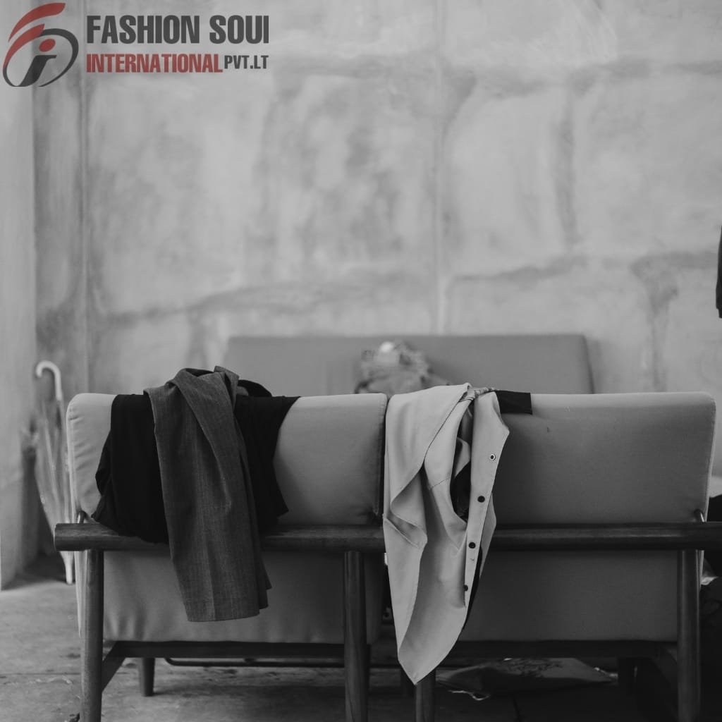 A black-and-white photo of a concrete-walled room with a sofa, on which various clothes are casually draped, and a wall logo reading "FASHION SOUL INTERNATIONAL PVT. LTD."