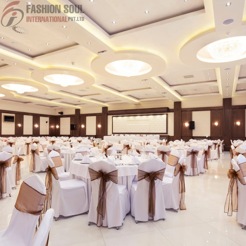An elegant banquet hall decorated for a formal event. The hall features a high ceiling with recessed lighting and large, oval-shaped chandeliers. The room is filled with round tables covered in white tablecloths, each surrounded by white chairs draped with white covers and tied with brown sheer sashes. The walls have dark wood paneling with soft lighting, and at the far end, there is a stage with a white backdrop. The overall ambiance is sophisticated and welcoming.