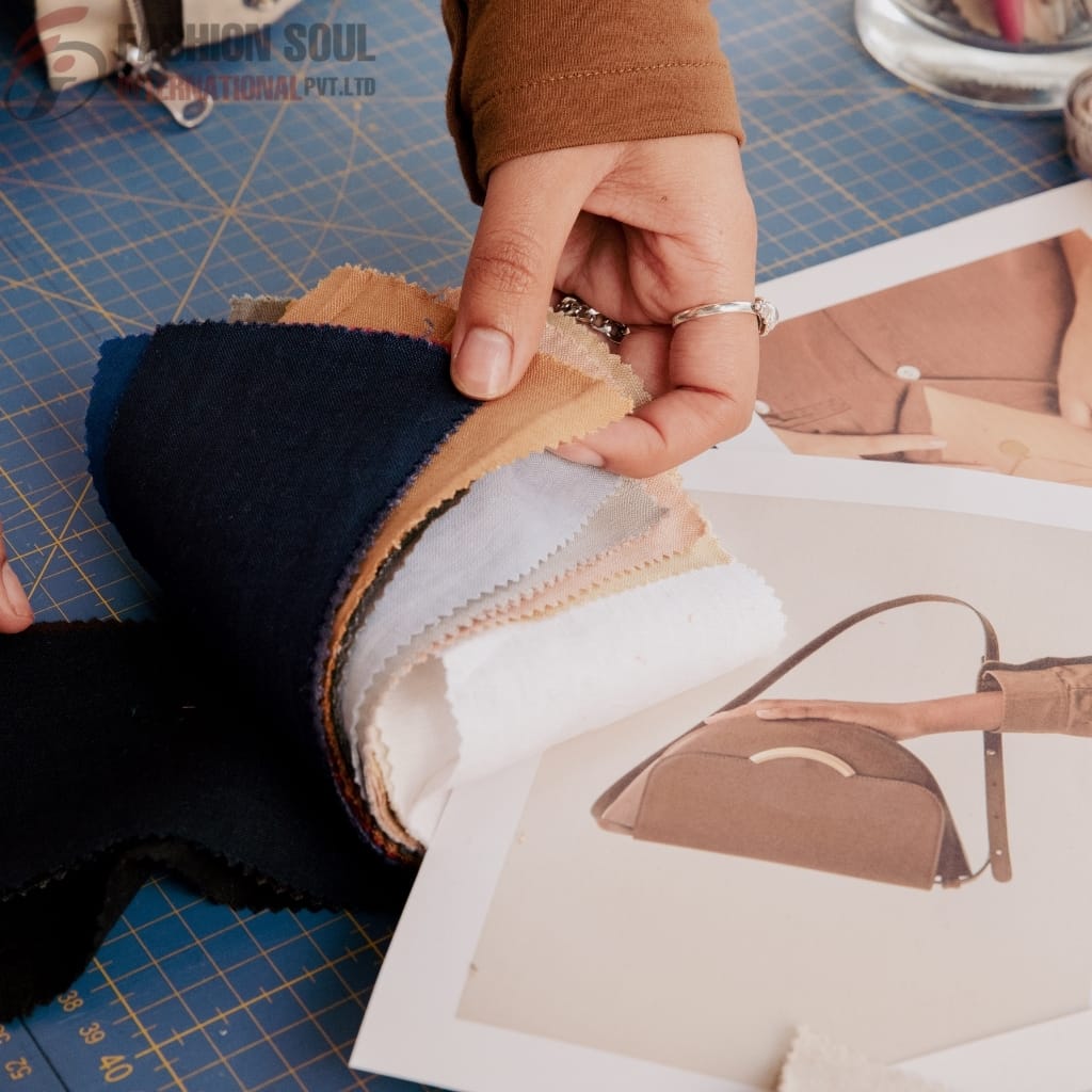 A person is holding a stack of fabric swatches in various colors, including navy, beige, light gray, white, and black, while working on a fashion or design project. The fabric swatches are placed on a cutting mat, with a printed image of a handbag and a model's outfit visible beneath the swatches.