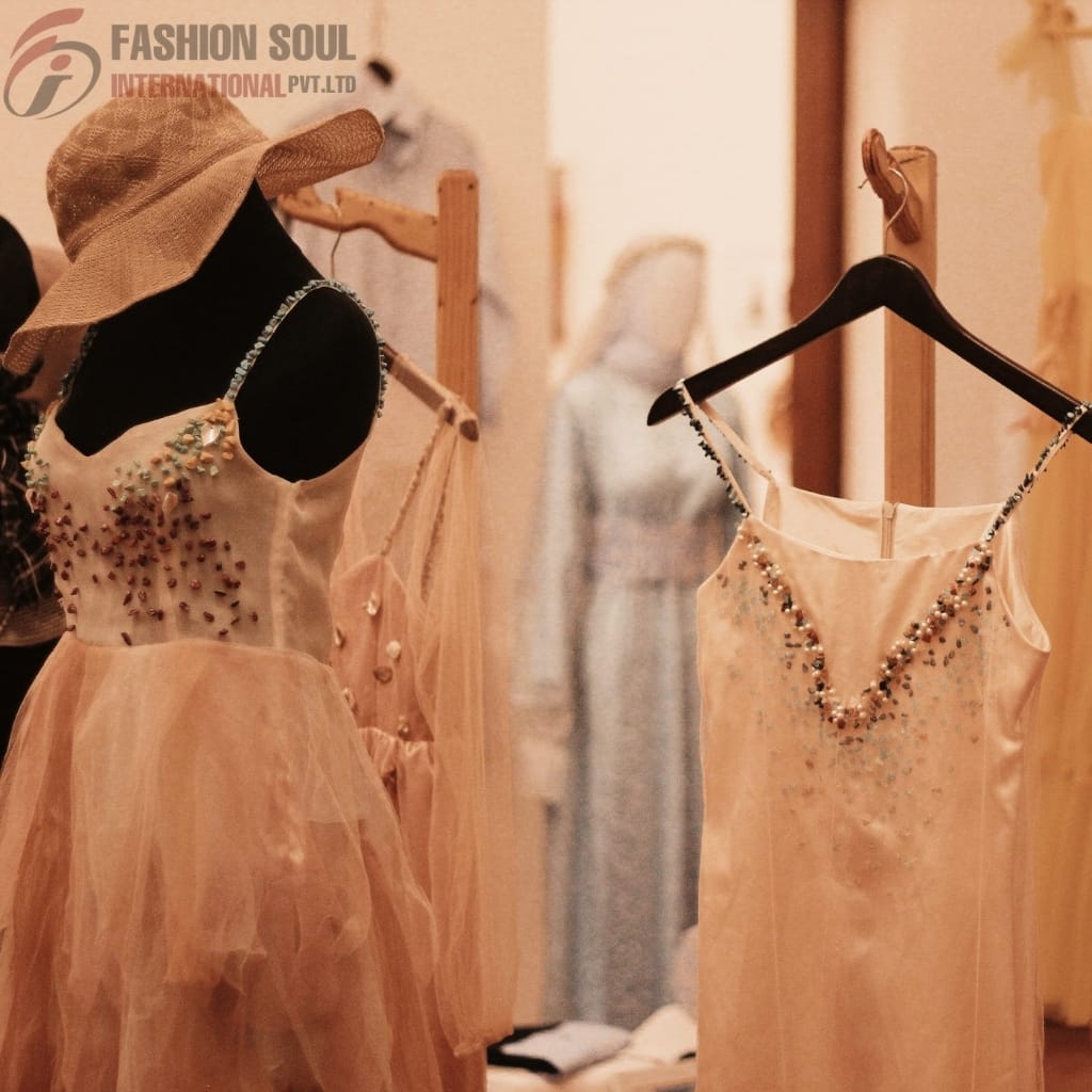 Two elegant peach-colored dresses with beaded embellishments displayed on mannequins and hangers inside a boutique. The dress on the left features a tulle skirt and a sleeveless top with floral beadwork, accompanied by a wide-brimmed straw hat. The dress on the right is a simple, sleeveless, beaded dress hung on a black hanger. In the background, there are other dresses on mannequins and clothing racks.