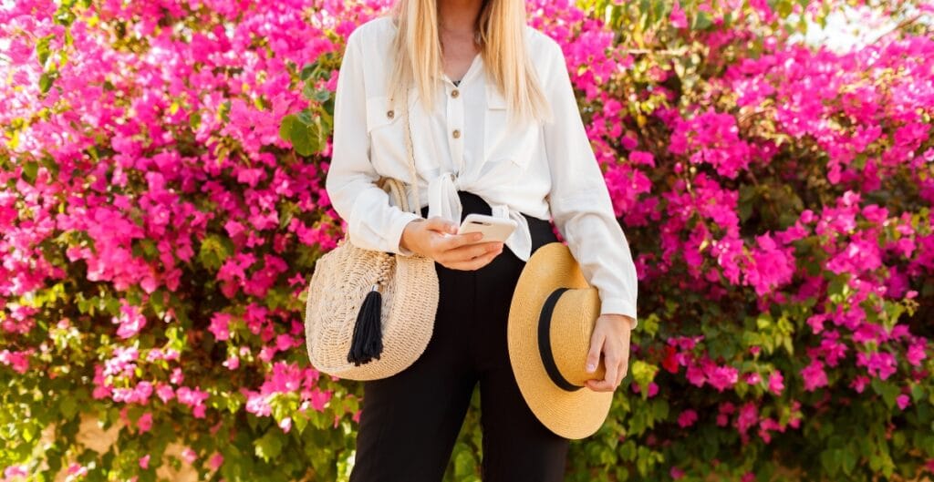 A woman standing in front of vibrant pink flowers, holding a smartphone in one hand and a straw hat in the other, with a woven bag hanging from her shoulder.
