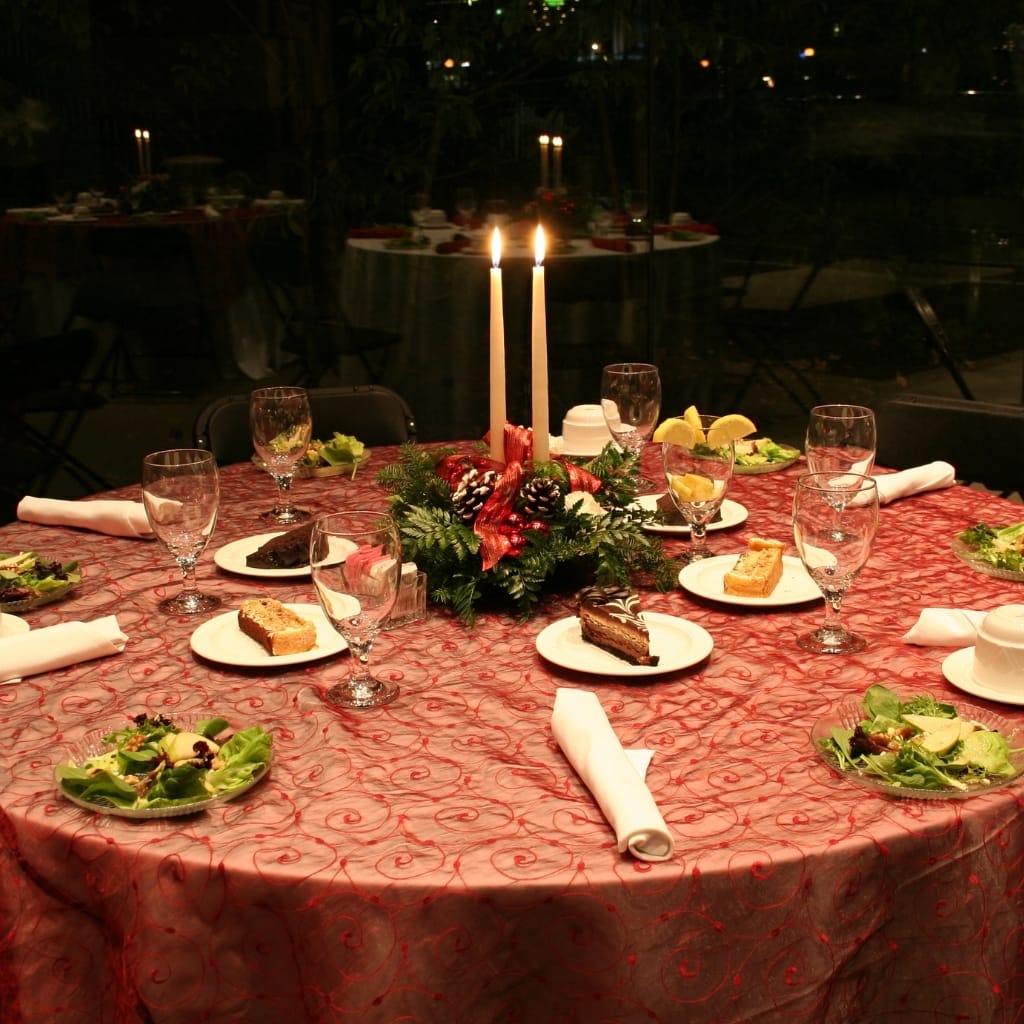 A beautifully decorated round dining table set for a formal dinner, featuring a red patterned tablecloth, candle centerpiece, salads, desserts, and neatly folded white napkins, with a softly lit background.