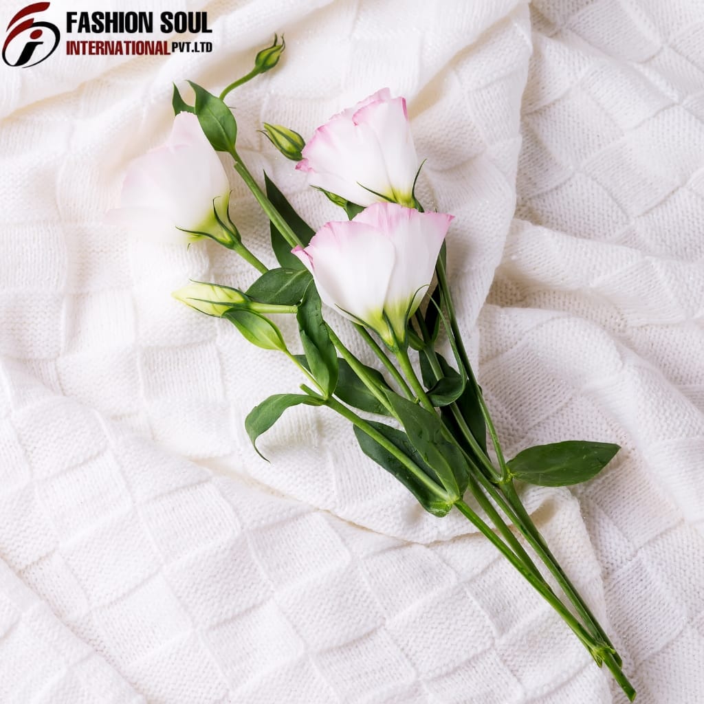 A small bouquet of white flowers with pink edges, resting on a white textured fabric background, accompanied by green leaves and buds.