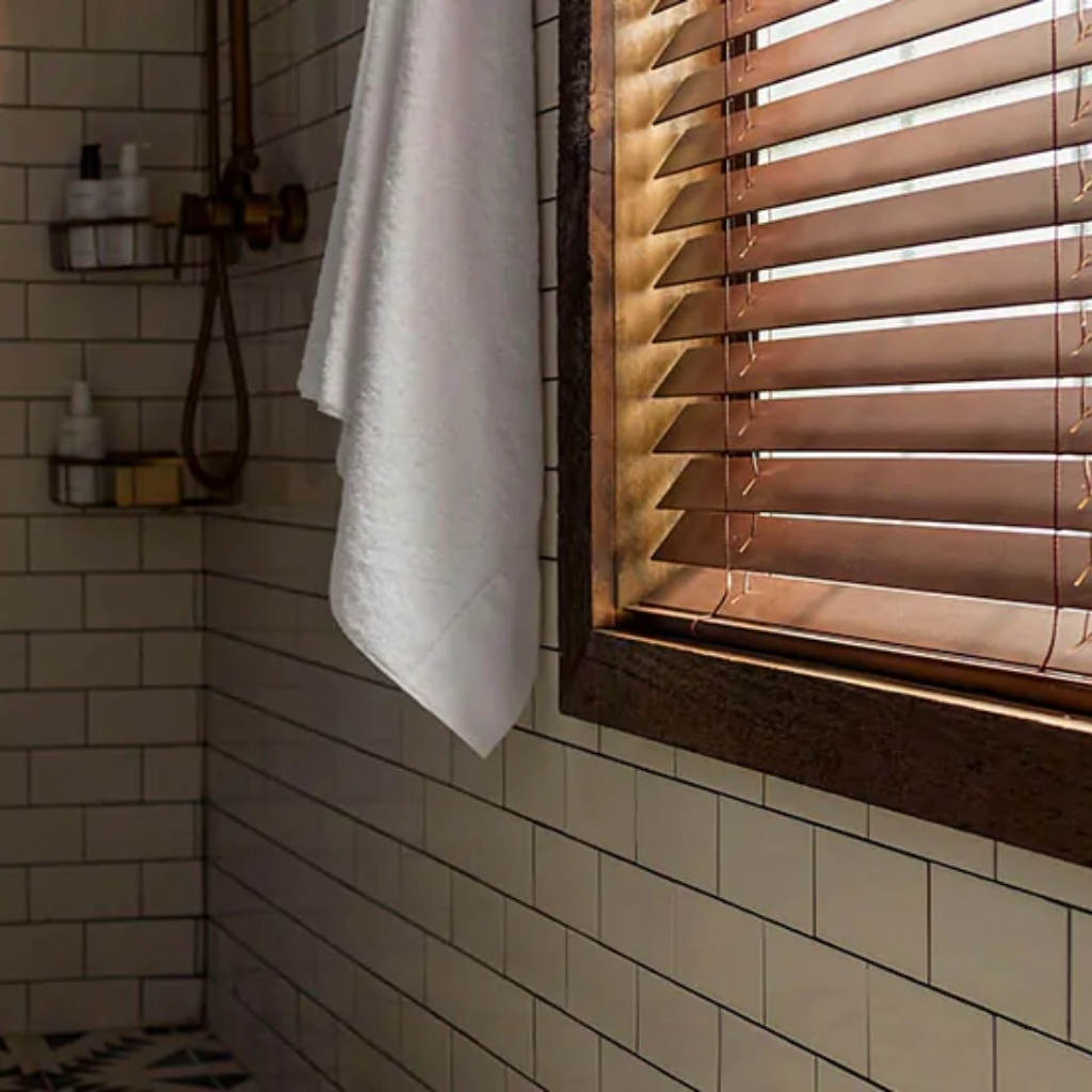 Close-up of a bathroom corner with beige tiled walls, a white towel hanging, and wooden window blinds.