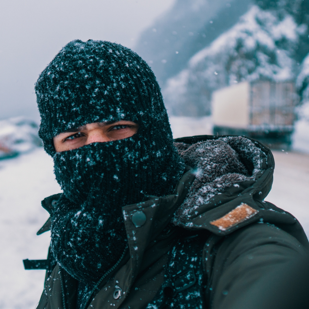 A person wearing a black knitted hat and a balaclava, bundled up against the cold, in a snowy outdoor setting.