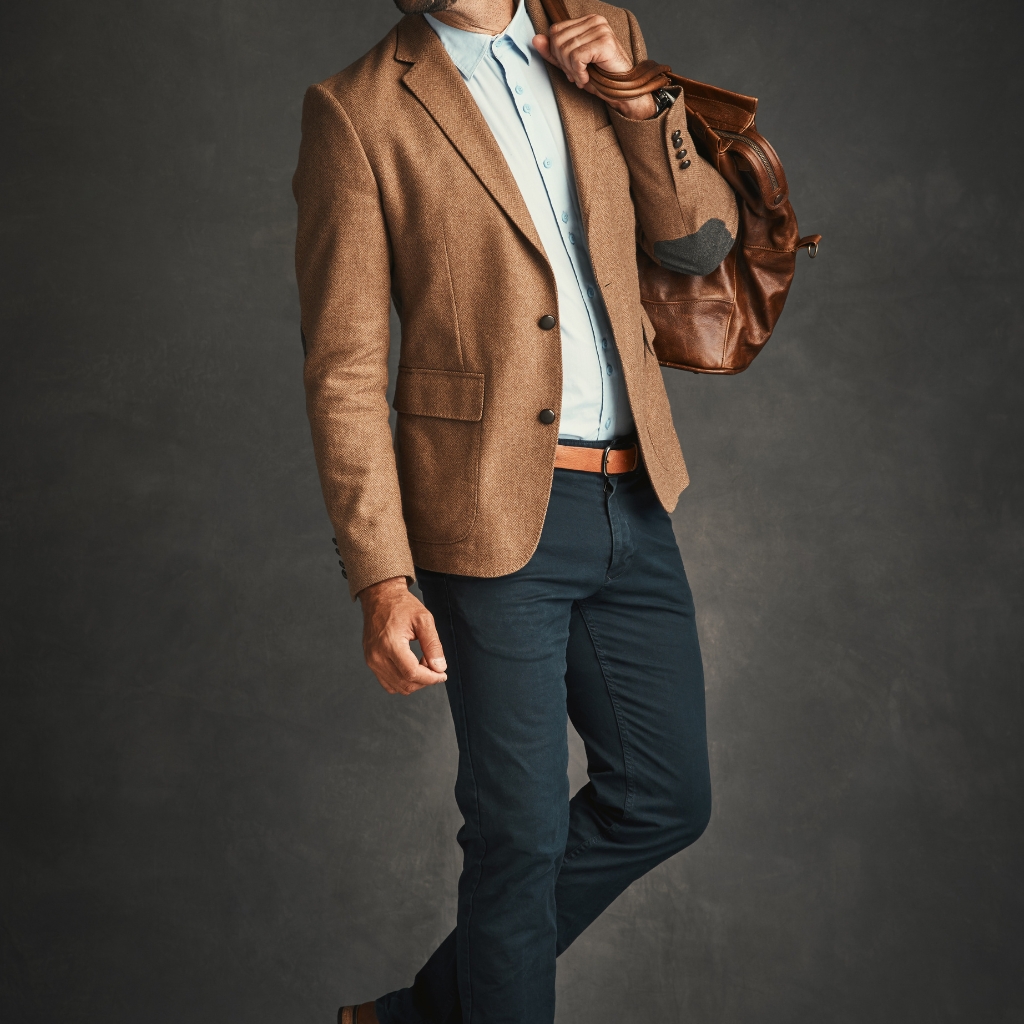 A man dressed in a stylish, semi-formal outfit featuring a brown sport coat, light blue shirt, dark jeans, and carrying a brown leather bag, standing against a dark background.