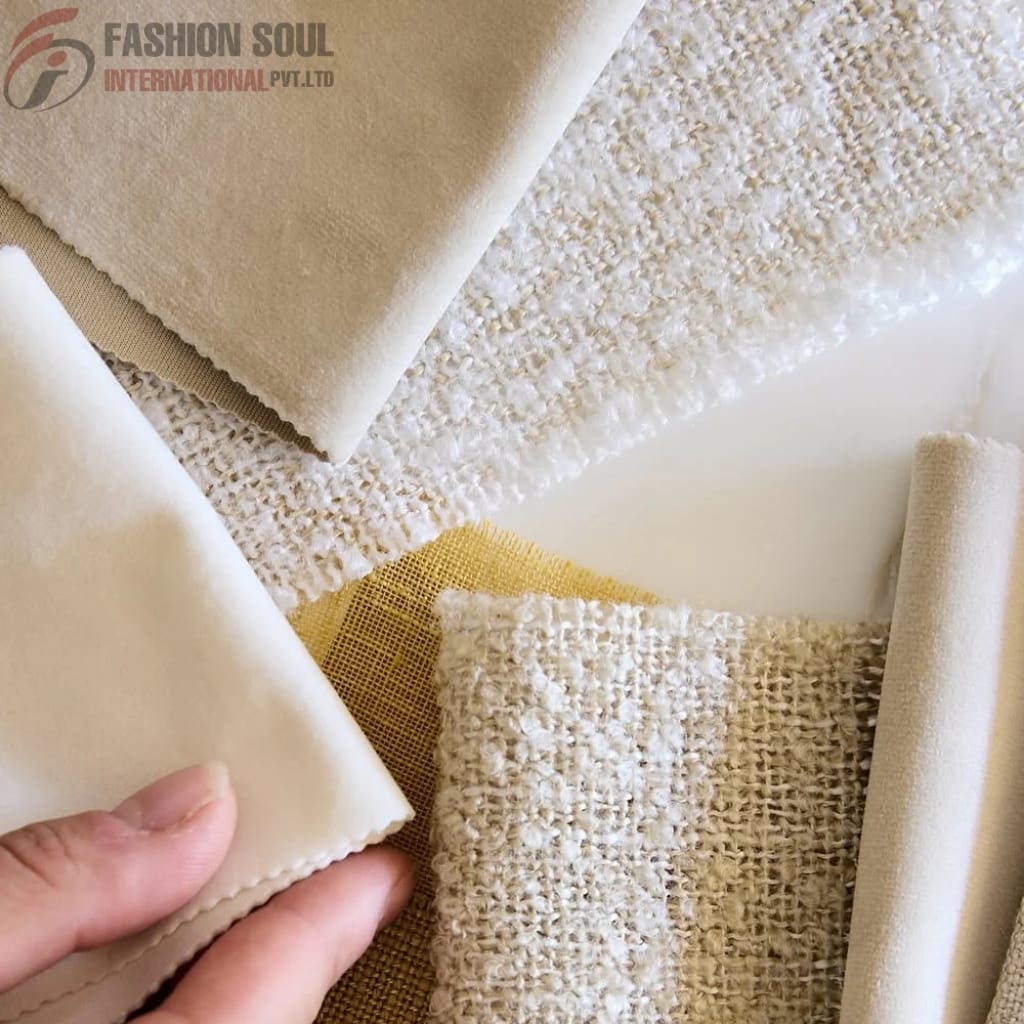 A close-up image showing a variety of fabric swatches in neutral tones, including smooth, textured, and woven materials. A person's hand is holding one of the fabric swatches in the lower left corner, and the fabric samples are arranged overlapping on a flat surface.