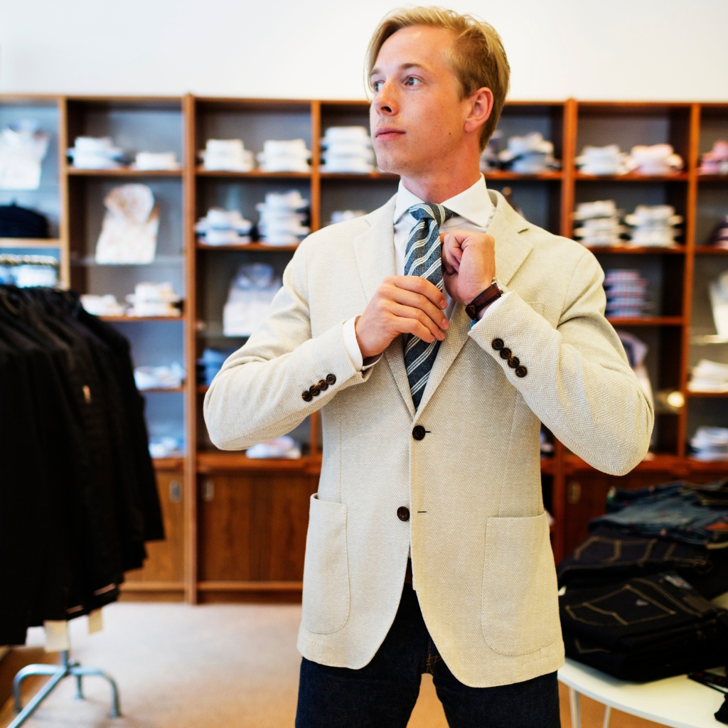 A young man in a light-colored blazer adjusting his tie while standing in a clothing store with shelves of folded shirts and pants in the background.