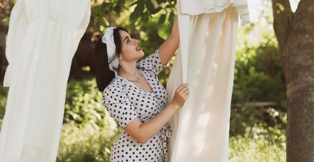 A woman with dark hair, wearing a polka-dot dress and a headscarf, looks up and smiles while hanging laundry on a clothesline outdoors surrounded by greenery.