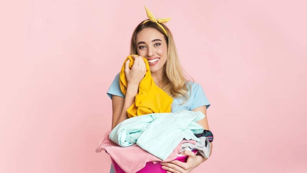 A smiling woman with a yellow headband holds a heap of colorful clothes against a pink background.