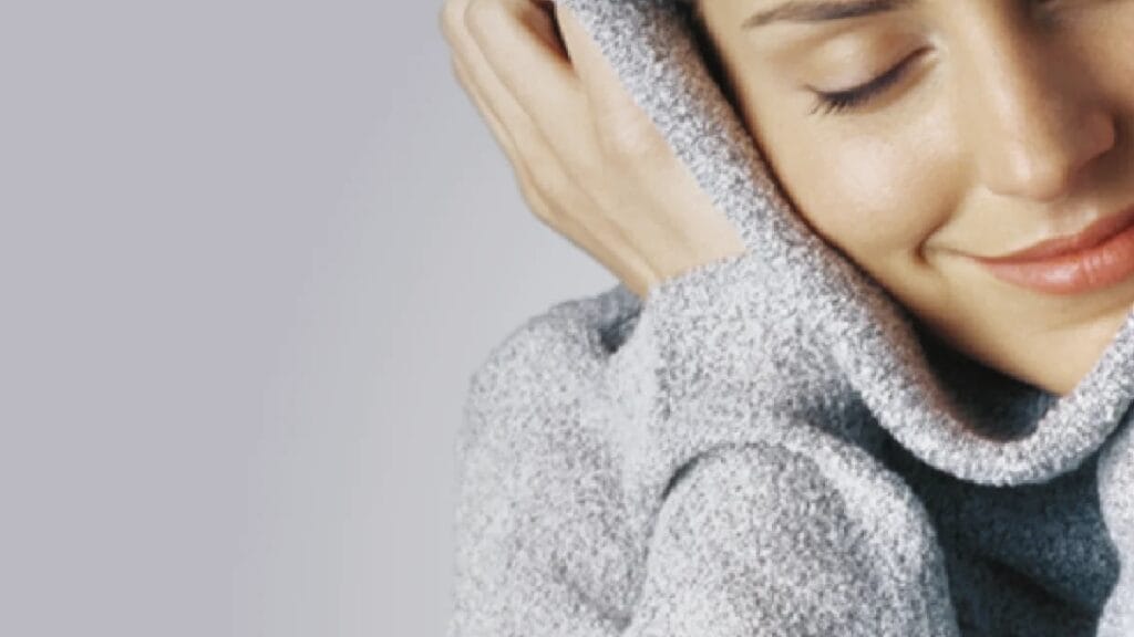 A woman with a soft smile rests her face in a gray knitted scarf, looking relaxed.
