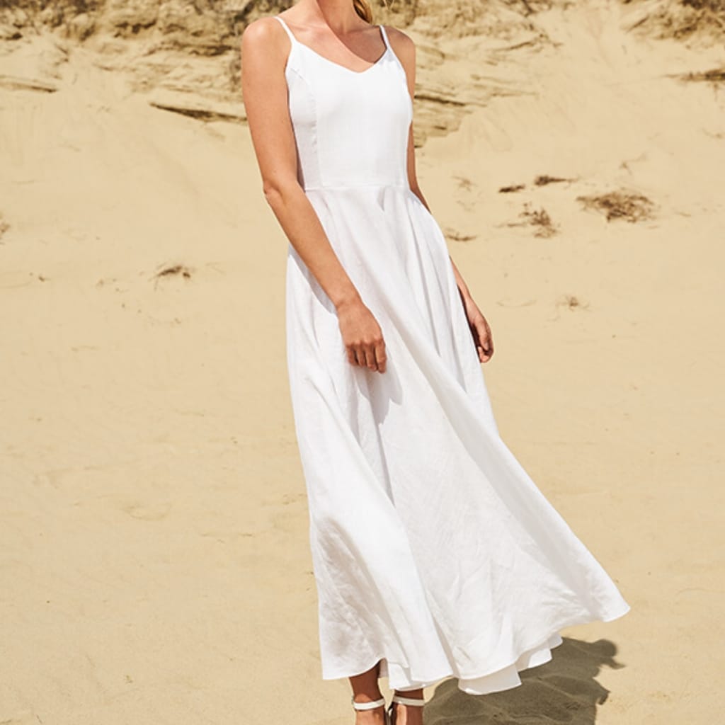 A woman wearing a flowing white sundress with thin straps, standing on a sandy beach with a rocky background.