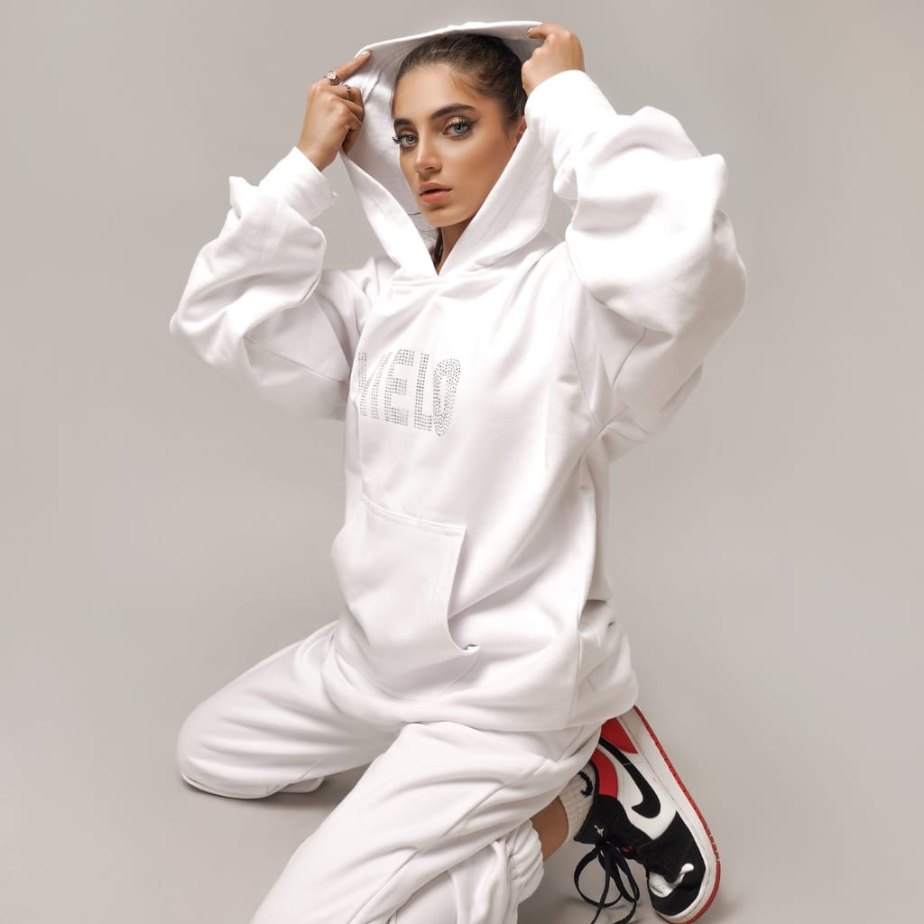 A young woman with styled hair and makeup is kneeling on the floor against a neutral background, wearing a white oversized hoodie and matching sweatpants, paired with black and white athletic shoes with red accents.