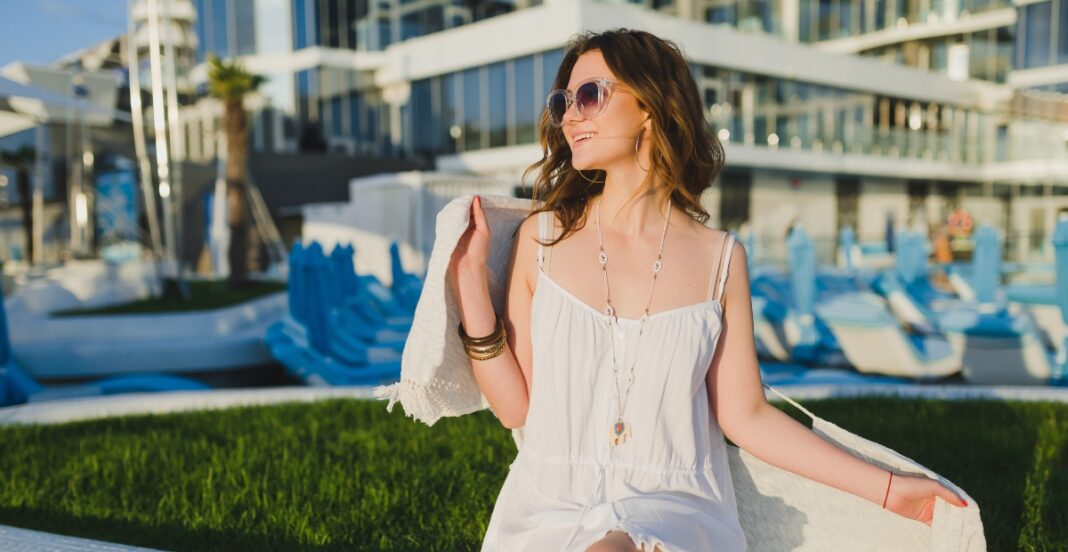 A woman with wavy brown hair and sunglasses is sitting outdoors by a marina, wearing a white dress and accessorized with necklaces and bracelets, holding a towel over her shoulder and smiling.