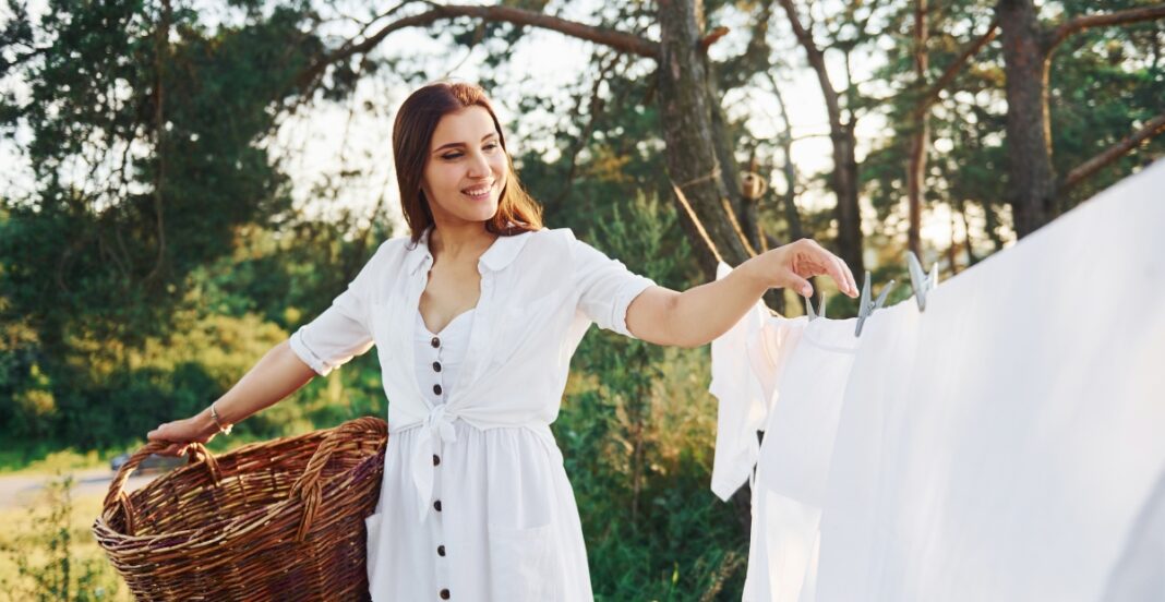 A woman in a white dress hanging laundry on a clothesline outdoors, holding a basket with one hand and using the other to clip clothes on the line, with trees and sunlight in the background.