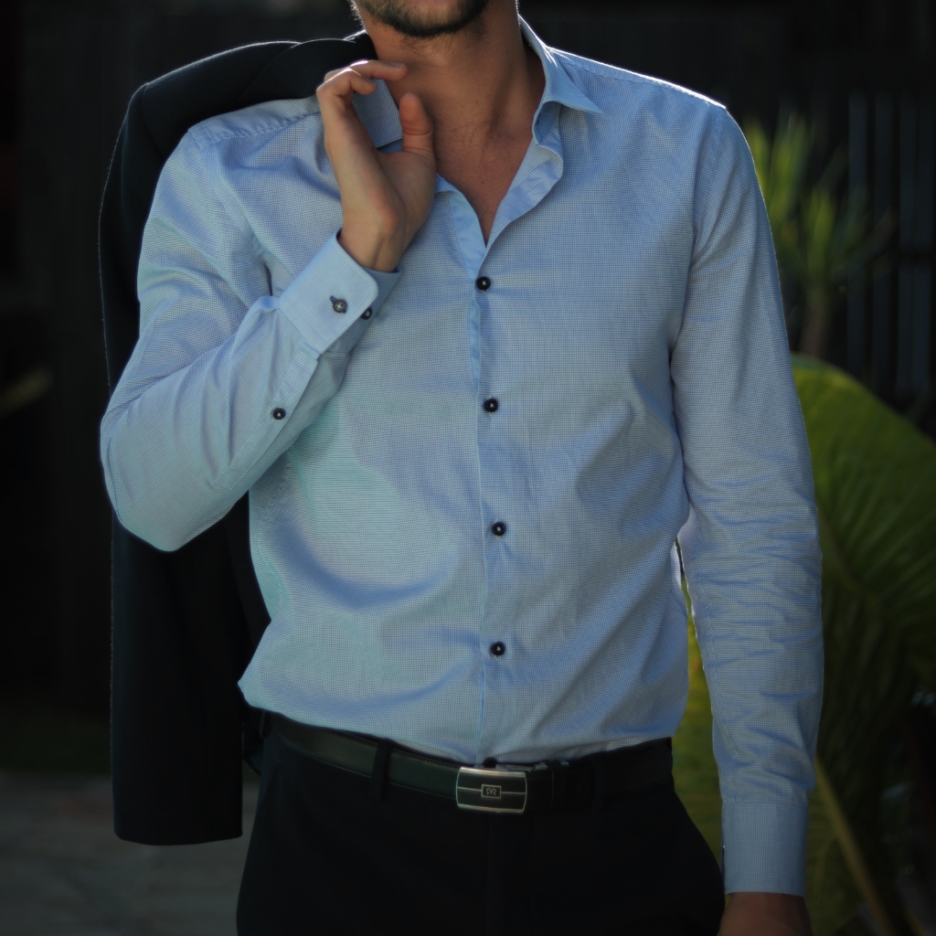 A man wearing a light blue, long-sleeved dress shirt with a subtle checkered pattern, unbuttoned at the collar, and black buttons. He is holding a dark suit jacket over his shoulder with one hand and is dressed in black pants with a black belt. The background appears to be outdoors with greenery.