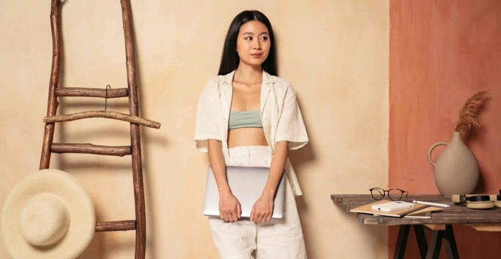 A young woman with long black hair, wearing a light green crop top and white outfit, stands against a textured beige and terracotta wall, holding a silver laptop. She is positioned near a rustic wooden ladder, a wide-brimmed straw hat, and a table with eyeglasses, a notebook, and a vase with dried grass.