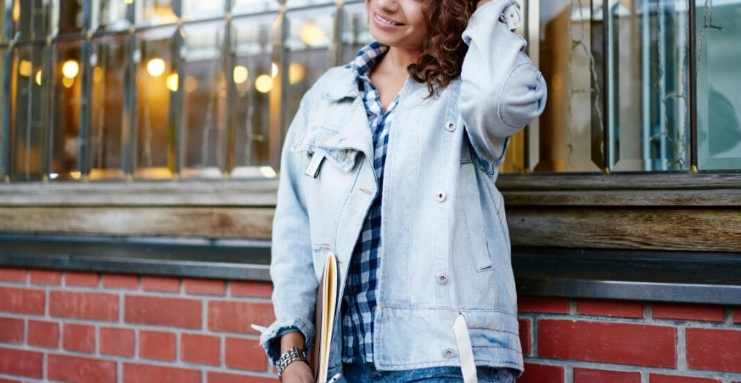 A young woman with curly hair smiling and standing outdoors against a brick wall with large windows behind her. She is wearing a light denim jacket over a blue checkered shirt and holding a notebook or folder.