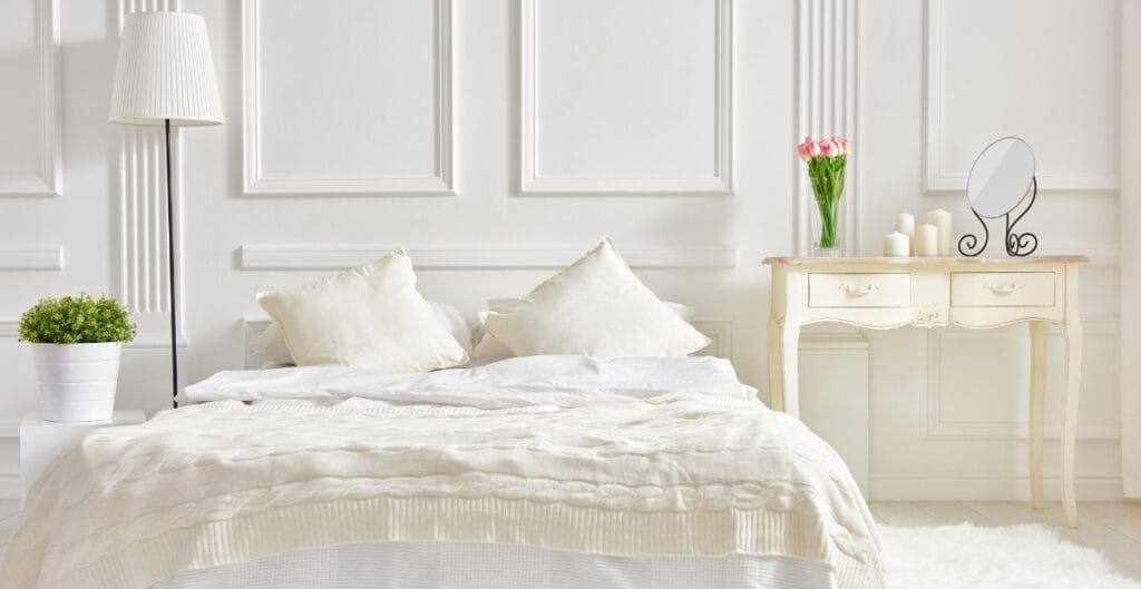 A bright, elegant bedroom with white paneled walls, a neatly made bed with white bedding and pillows, a tall white floor lamp on the left, a small white nightstand with a green potted plant, and a cream-colored dresser with pink flowers and candles on top, along with a small round mirror.